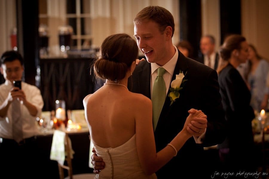 carolina inn weddings | allison and matthew A bride and groom share a first dance in a warmly lit reception hall. The groom is dressed in a suit with a light green tie, and the bride wears a strapless white gown. Guests are visible in the background, some capturing the moment with cameras.