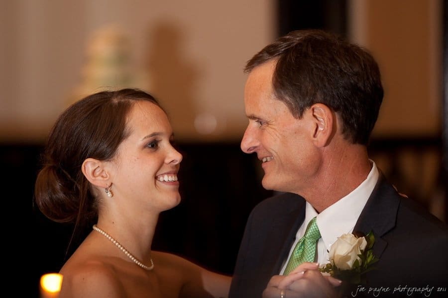 carolina inn weddings | allison and matthew A woman and a man are dancing together, smiling at each other. She is wearing a pearl necklace and earrings, and he is in a suit with a green tie and a white rose boutonniere. The background is softly blurred.