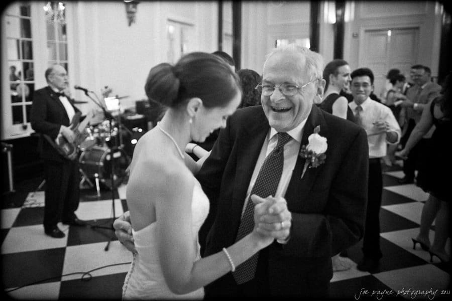 carolina inn weddings | allison and matthew A young woman in a wedding dress dances joyfully with an elderly man in a suit and tie at a formal event. A band plays instruments in the background. The black and white image captures the lively, celebratory atmosphere.