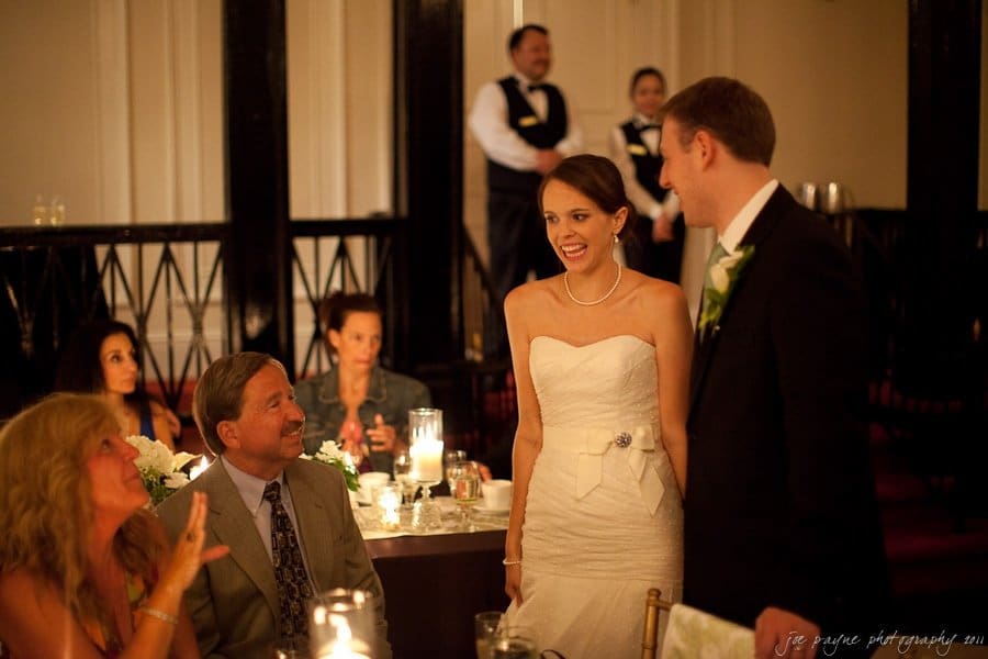 carolina inn weddings | allison and matthew A bride and groom are standing and smiling at wedding guests seated at a table. The bride is wearing a white strapless gown, and the groom is in a suit. The room is warmly lit with candles and decorations.