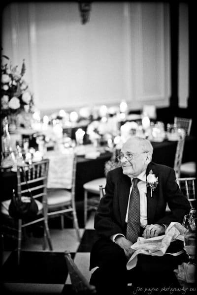 carolina inn weddings | allison and matthew Black and white image of an elderly man in a suit sitting at a table decorated for an event. The background has elegantly set tables with floral arrangements and candles.