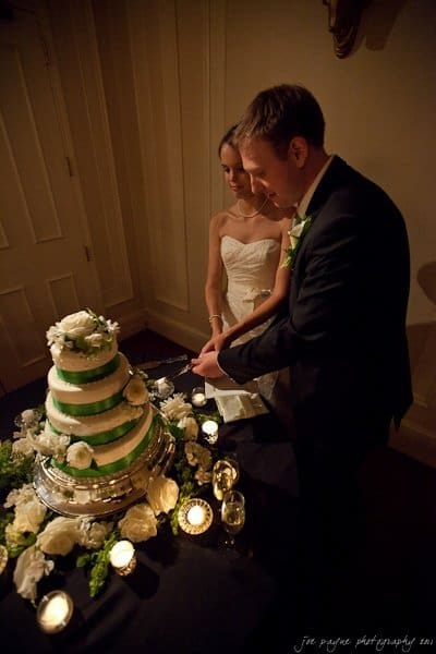 carolina inn weddings | allison and matthew A bride and groom stand together as they cut a tiered wedding cake adorned with white flowers and green accents. The table is decorated with flowers and candles. The room is softly lit, creating a warm ambiance.
