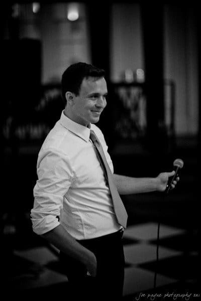 carolina inn weddings | allison and matthew Man in a dress shirt and tie holding a microphone, smiling, captured in black and white. The background features a blurred interior setting with faint lights.