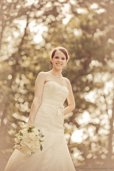 carolina inn weddings | allison and matthew A bride in a strapless white wedding gown holds a bouquet of white flowers. Sunlight filters through the trees in the background, creating a soft, romantic atmosphere.