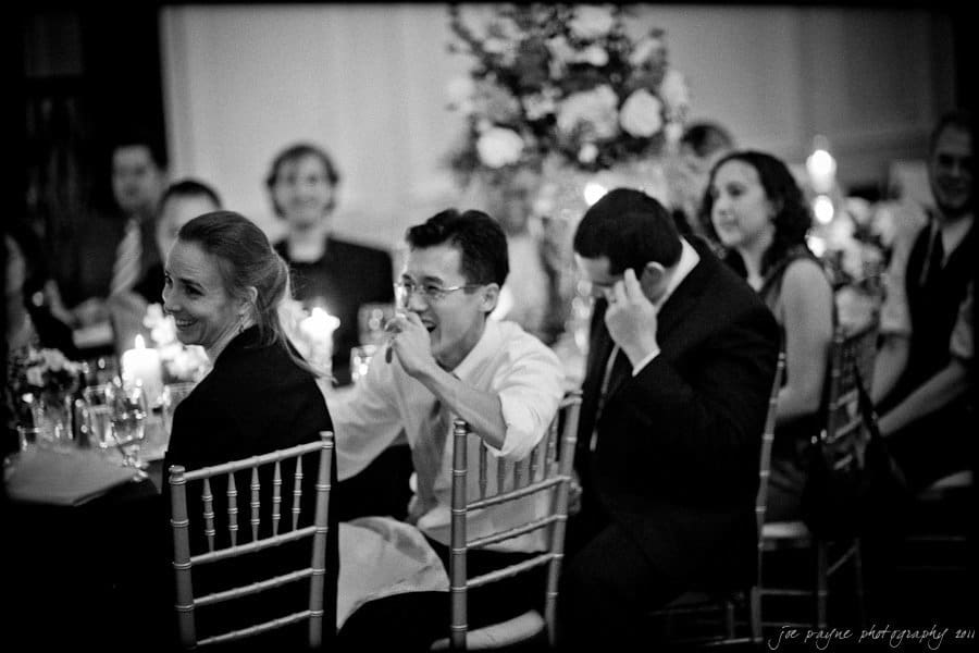 carolina inn weddings | allison and matthew Guests sitting at a wedding reception, laughing and enjoying themselves. The image is in black and white, capturing a lively atmosphere with people seated around a table decorated with floral arrangements and lit candles.