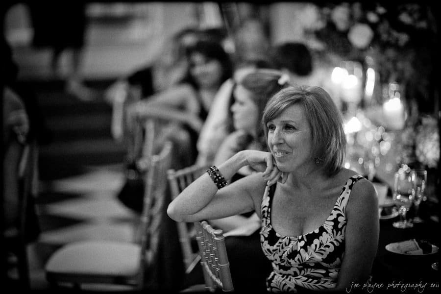 carolina inn weddings | allison and matthew A black and white photo of a woman seated at an event, smiling and resting her chin on her hand. She is wearing a patterned dress and a bracelet, with a background of guests and candlelit tables.
