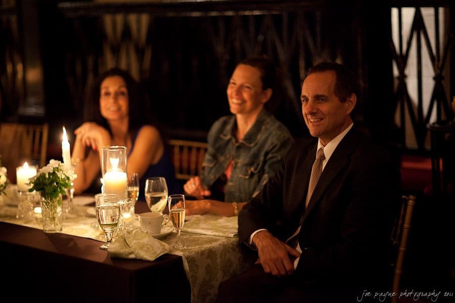 carolina inn weddings | allison and matthew Three people sit at a candlelit table in an elegant setting. They are smiling and appear to be enjoying themselves. Two women are on the left and a man in a suit is on the right. The table is decorated with flowers and elegant tableware.