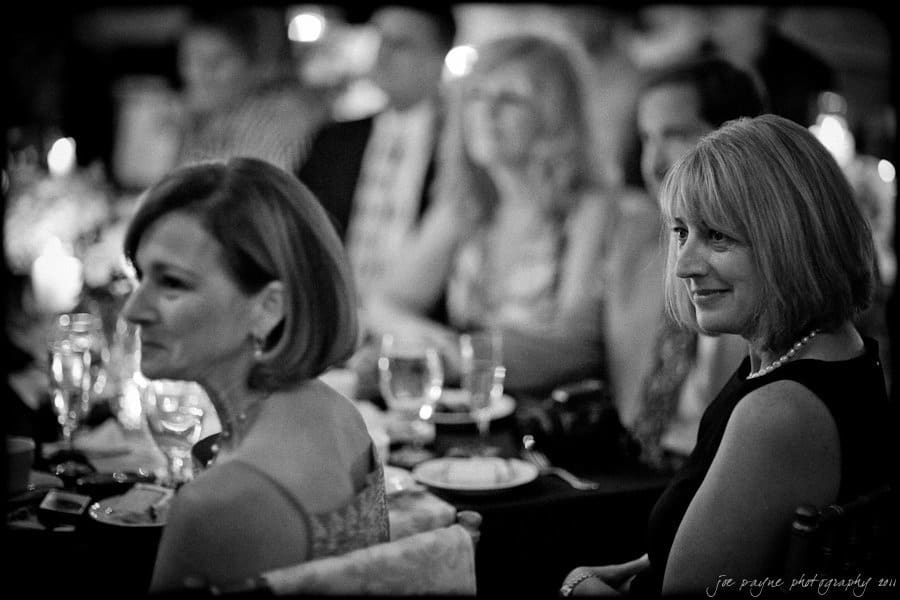 carolina inn weddings | allison and matthew Two women at a dinner event, seated at a table with glasses and tableware. Both are looking towards the front, with soft lighting creating a warm atmosphere. A blurred background shows other attendees.