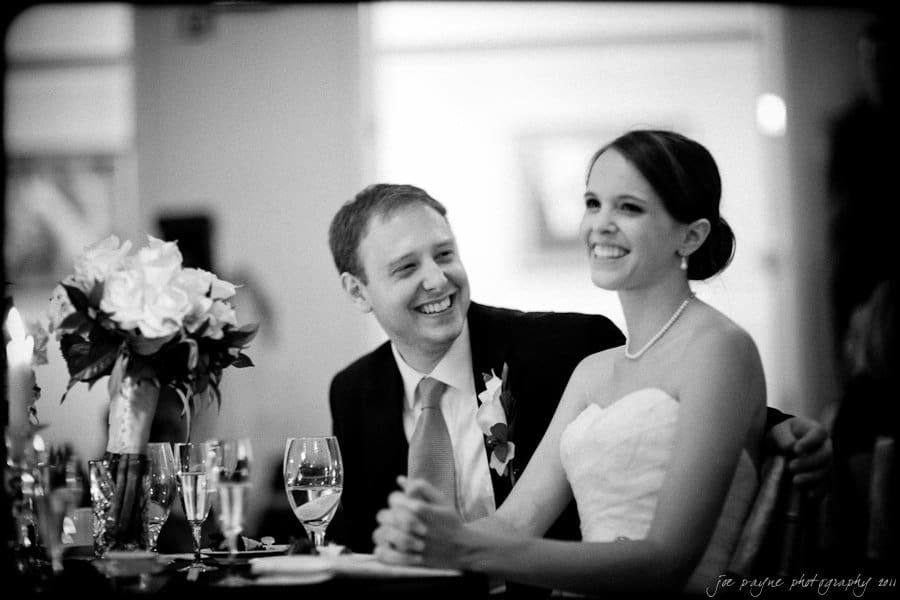 carolina inn weddings | allison and matthew A bride and groom sit together at a wedding reception, smiling and looking towards each other. The bride is wearing a strapless dress with a pearl necklace, and the groom is in a suit with a boutonniere. A floral centerpiece is visible on the table.