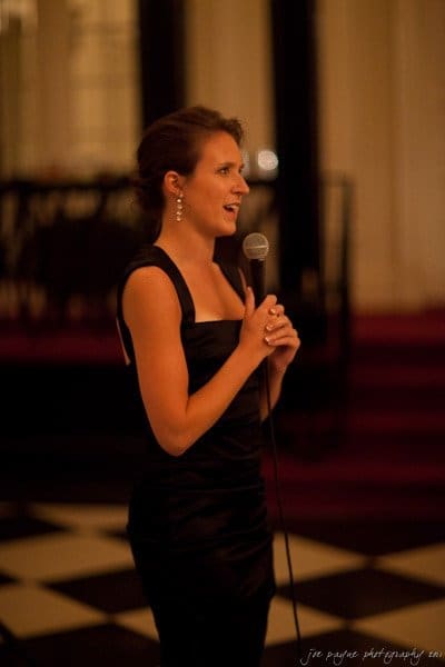 carolina inn weddings | allison and matthew A woman in a black evening dress speaks into a microphone. She stands on a black-and-white checkered floor with a blurred background, possibly a formal event or gathering.