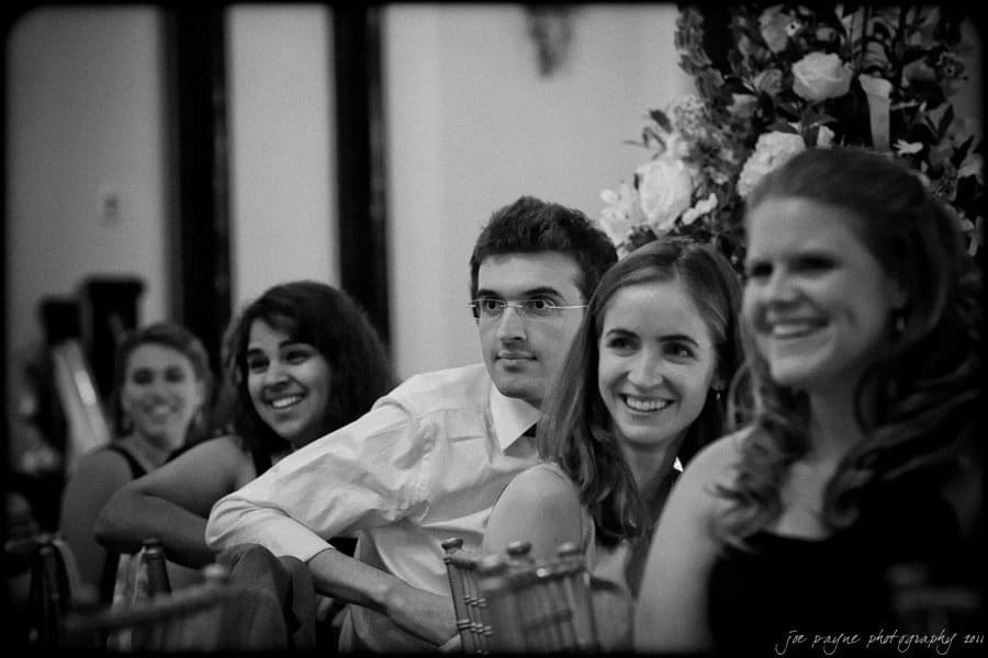 carolina inn weddings | allison and matthew A group of people sitting closely together, smiling and appearing engaged, in a candid black-and-white photograph. They are seated in a formal setting with floral decorations visible in the background.