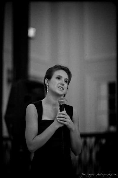 carolina inn weddings | allison and matthew A person in a dark dress holds a microphone, speaking or singing. The setting appears to be indoors with a soft focus background. The image is in black and white, adding a classic, timeless feel.