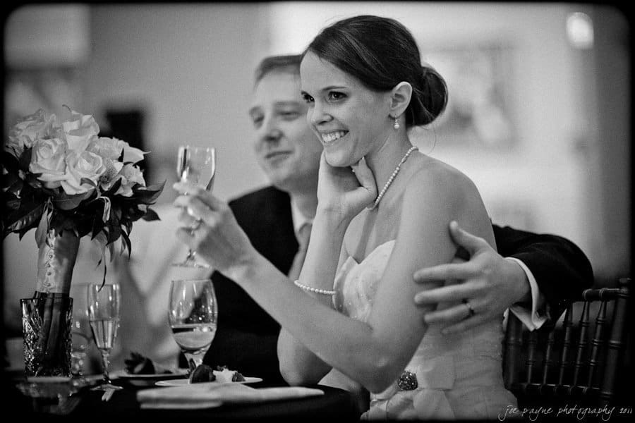 carolina inn weddings | allison and matthew A bride and groom sitting together at a reception. The bride smiles while holding a wine glass, and the groom sits beside her with his arm around her. A bouquet of flowers and a piece of cake are on the table in front of them.