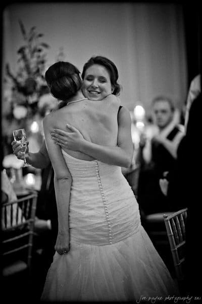 carolina inn weddings | allison and matthew Two women in wedding dresses embrace warmly at a reception. One holds a champagne glass. The background features guests seated at tables with floral decorations and candlelight, creating an elegant and celebratory atmosphere.