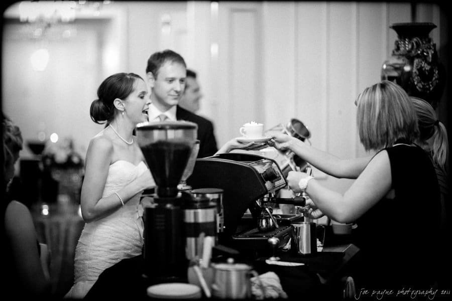 carolina inn weddings | allison and matthew A bride in a wedding dress joyfully accepts a coffee drink from a barista at a counter. Other guests and another barista are visible in the background. The scene is set in a formal indoor venue.