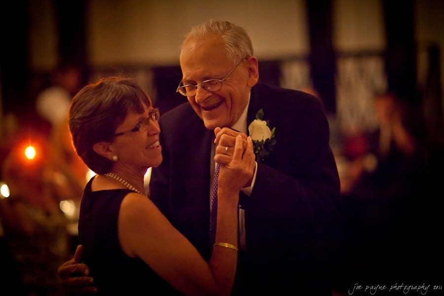 carolina inn weddings | allison and matthew An elderly couple, smiling and holding hands, dances together in a warmly lit room. He wears a dark suit with a white rose boutonniere, and she is dressed in a sleeveless top and pearls. The background features soft, blurred lights.