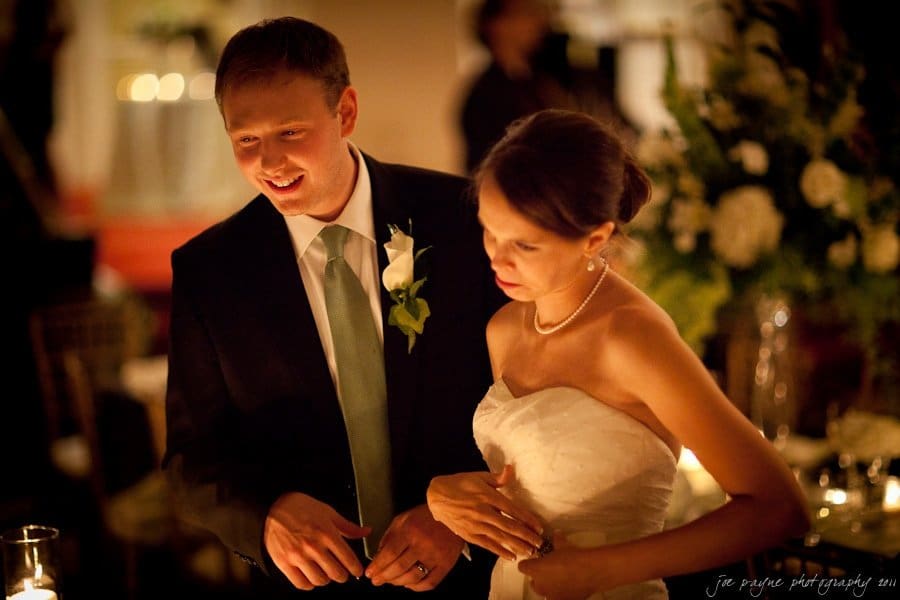 carolina inn weddings | allison and matthew A bride and groom smile warmly as they stand together at their wedding reception. The groom wears a dark suit with a light tie and boutonniere, while the bride is in a white strapless gown. Soft lighting and floral decorations create an intimate setting.