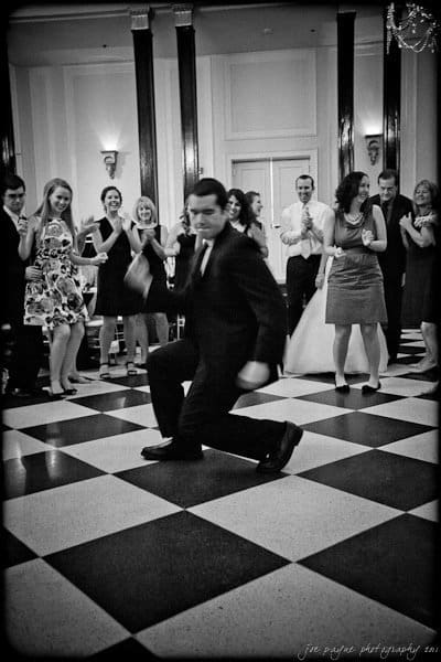 carolina inn weddings | allison and matthew A man in a suit energetically dances on a checkered floor at an indoor event, surrounded by people clapping and smiling. The background features tall columns and wall sconces, creating a festive atmosphere.