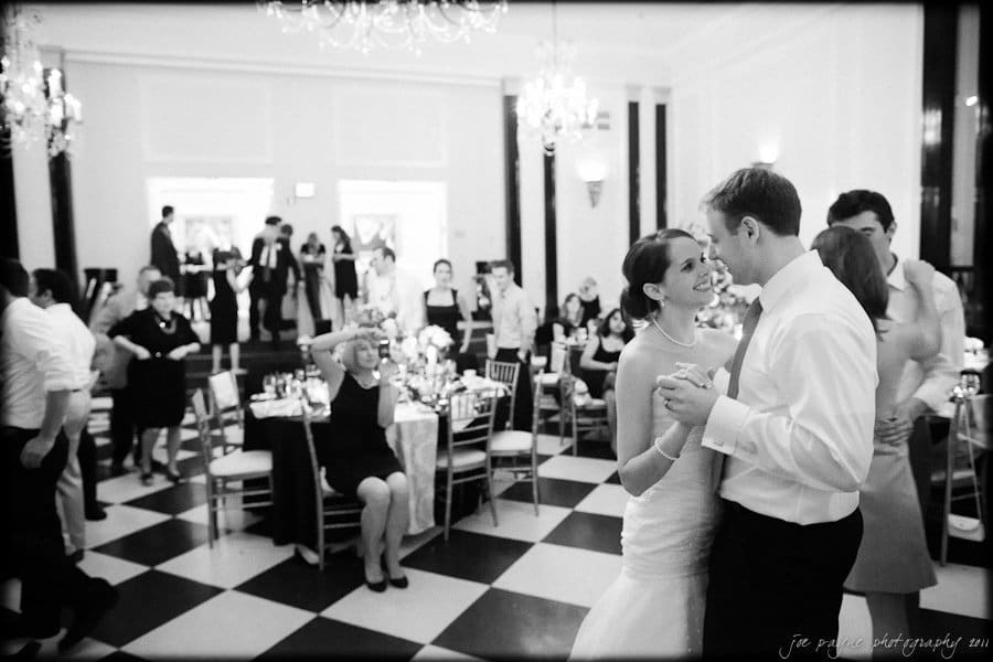 carolina inn weddings | allison and matthew A black and white photo of a couple dancing at a formal event. They are surrounded by guests seated at tables and standing in the background. The room has elegant chandeliers and checkered flooring.
