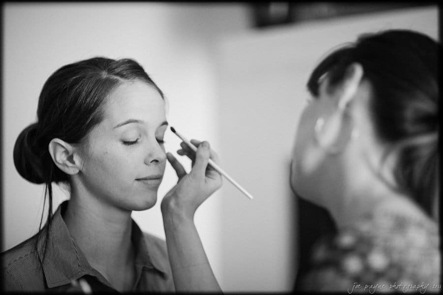 carolina inn weddings | allison and matthew A black and white image of a woman having her makeup done. She has her eyes closed while another woman stands in front, holding a makeup brush near her eye. The setting appears calm and intimate.