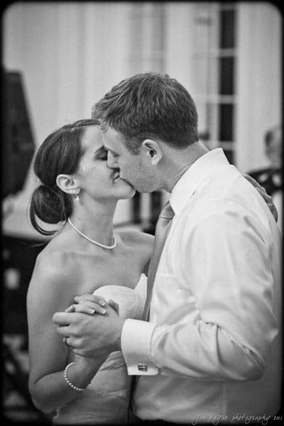 carolina inn weddings | allison and matthew A bride and groom share a tender kiss while dancing at their wedding. The bride is wearing a strapless dress and pearl necklace, and the groom is in a shirt and tie. Both are holding hands, creating a romantic and intimate moment. The image is in black and white.