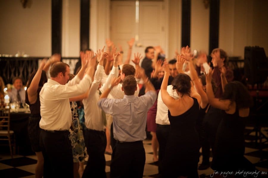 carolina inn weddings | allison and matthew A group of people forms a circle on a black-and-white checkered dance floor, raising their hands in celebration. They are dressed in formal attire, and the background features elegant decor and dim lighting, suggesting a festive occasion.