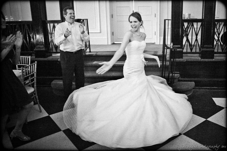 carolina inn weddings | allison and matthew A bride dances joyfully in a strapless wedding gown, spinning and smiling. A man claps and smiles at her. The setting is an elegant room with checkered flooring and white-paneled walls. The photo is in black and white.