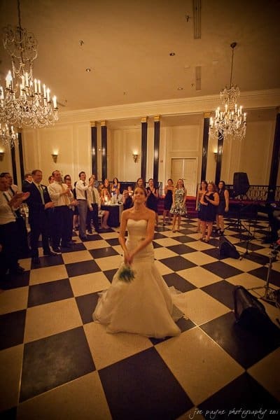 carolina inn weddings | allison and matthew A bride in a white gown stands with a bouquet in the center of a black and white checkered floor. Guests gather around the ballroom, smiling and clapping under elegant chandeliers.