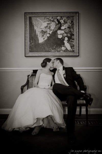 carolina inn weddings | allison and matthew A bride and groom share a kiss while sitting on a bench under a framed painting. The bride is in a strapless gown, and the groom is in a suit and tie. The scene is captured in black and white.