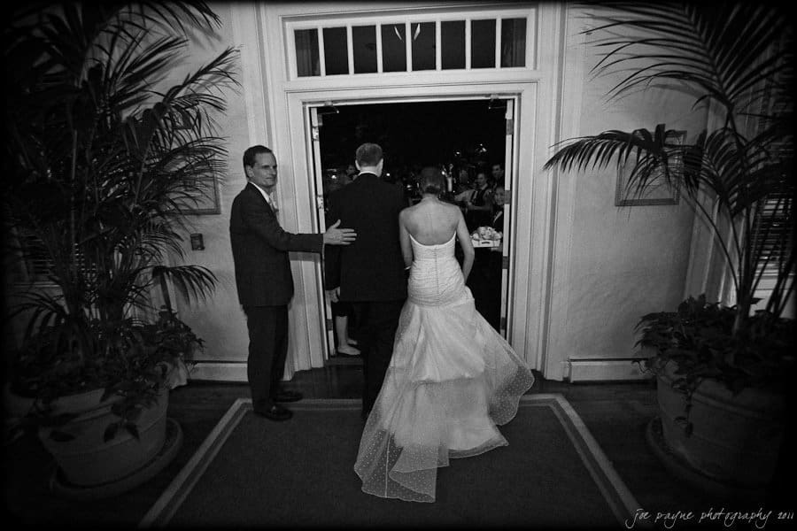 carolina inn weddings | allison and matthew A bride and groom, seen from behind, enter a doorway as a man gestures them forward. The bride wears a strapless gown, and the groom is in a dark suit. They are framed by large potted plants, and guests can be seen in the background.