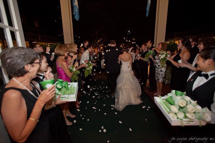 carolina inn weddings | allison and matthew A bride and groom walk down an aisle lined with cheering guests. The bride wears a white gown, and the groom is in a suit. Guests hold green cones and throw flower petals as the couple exits under a canopy.