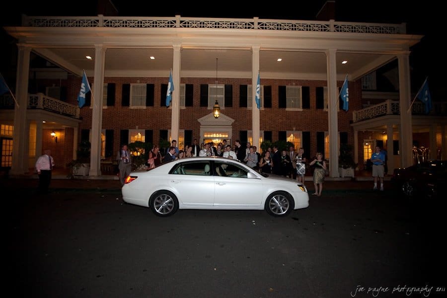 carolina inn weddings | allison and matthew A white sedan is parked in front of a large, well-lit building adorned with flags at night. A crowd of people stands on the buildings porch, many waving or taking photos, suggesting a celebratory event.