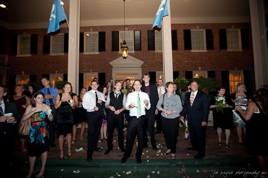 carolina inn weddings | allison and matthew A group of people dressed in formal attire stand outside a brick building with tall columns. Some are holding flowers, and theres a sense of celebration in the air. Blue flags hang above them.