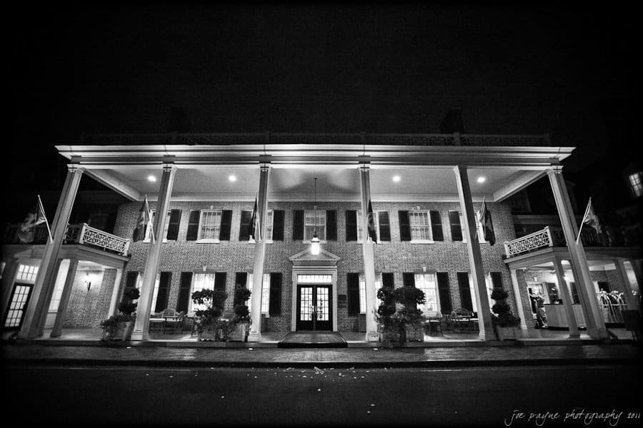 carolina inn weddings | allison and matthew Black and white photo of a stately brick building with large white columns and a lit portico. Flags adorn the front, and tables are visible on a porch. This elegant structure appears to be a historic inn or hotel.