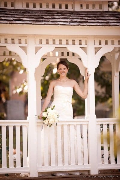 carolina inn weddings | allison and matthew A bride in a strapless white gown stands smiling inside a gazebo. She is holding a bouquet of white flowers and looking at the camera. The gazebo is white with decorative woodwork. Light filters softly through trees in the background.
