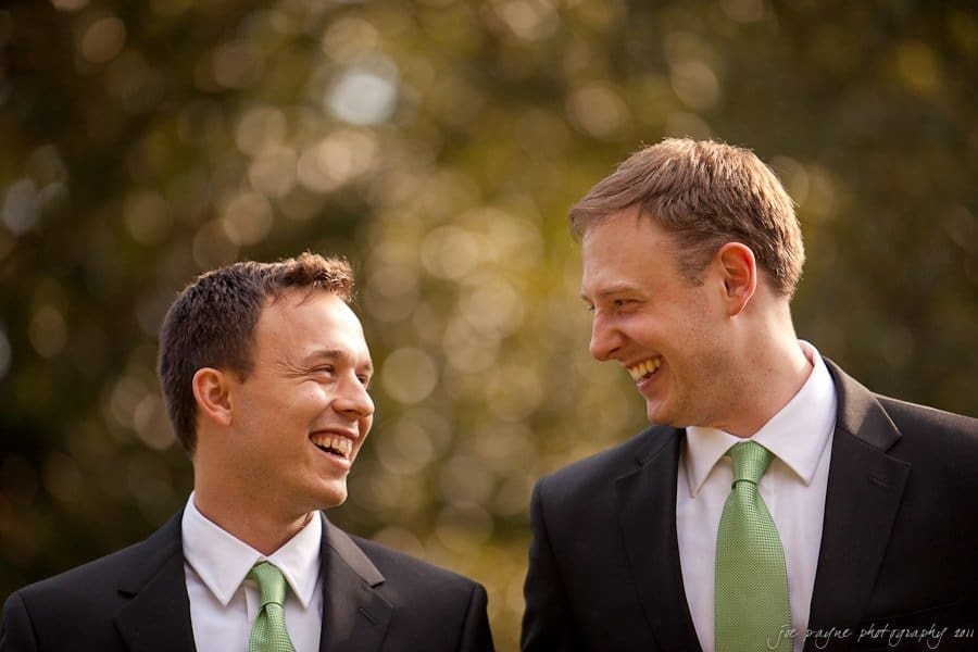 carolina inn weddings | allison and matthew Two men in suits with green ties are standing outdoors, facing each other and smiling. The background is softly blurred with greenery and light bokeh effects.