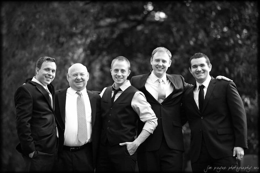 carolina inn weddings | allison and matthew Five men in suits and ties stand together outdoors, smiling at the camera. The background is blurred with greenery.