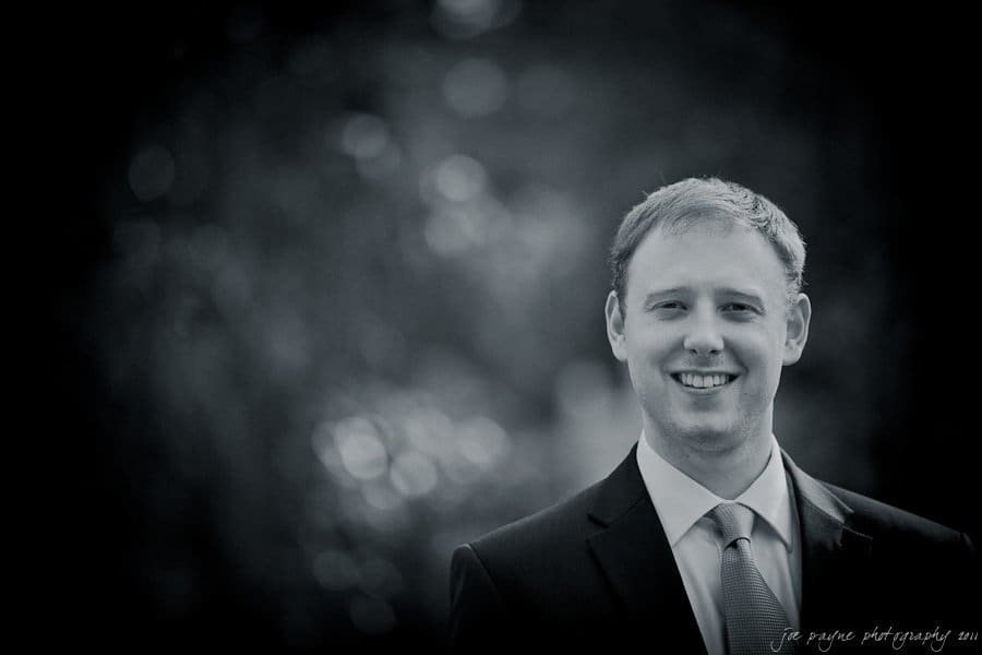 carolina inn weddings | allison and matthew A black and white portrait of a man in a suit and tie, smiling at the camera. The background is softly blurred with bokeh effects from trees.