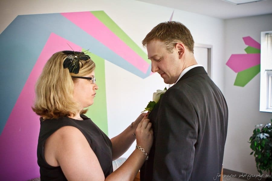 carolina inn weddings | allison and matthew A woman in a black dress adjusts a boutonniere on a mans suit. They are in a room with colorful geometric wall art and a small window in the background. The man looks down at the boutonniere.