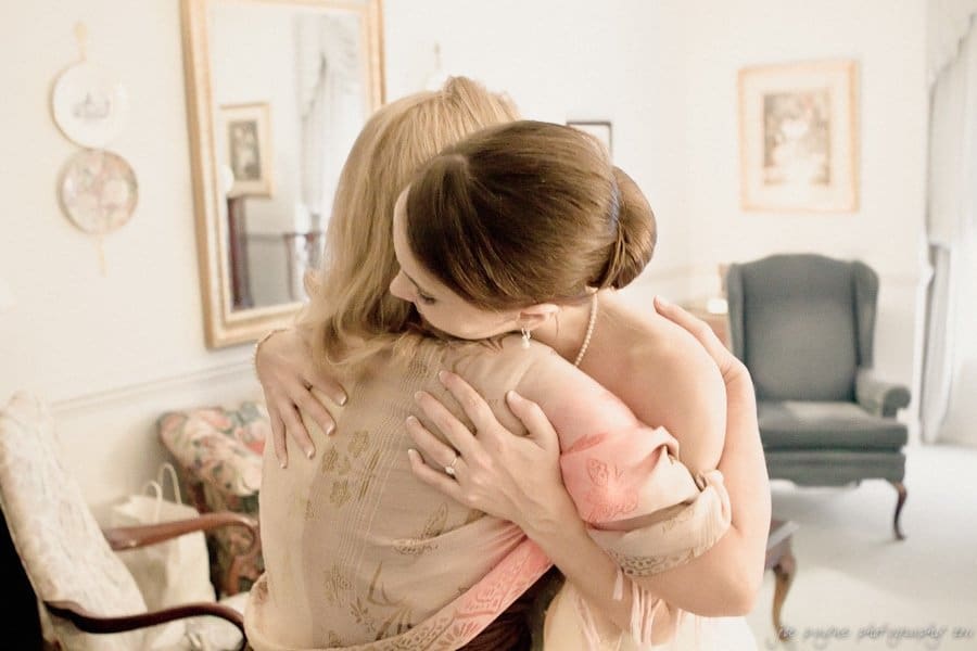 carolina inn weddings | allison and matthew Two women embracing in a warmly lit room. One wears a strapless dress and a pearl necklace, while the other wears a patterned shawl. The room is decorated with framed artwork and a dark armchair. The mood is intimate and heartfelt.