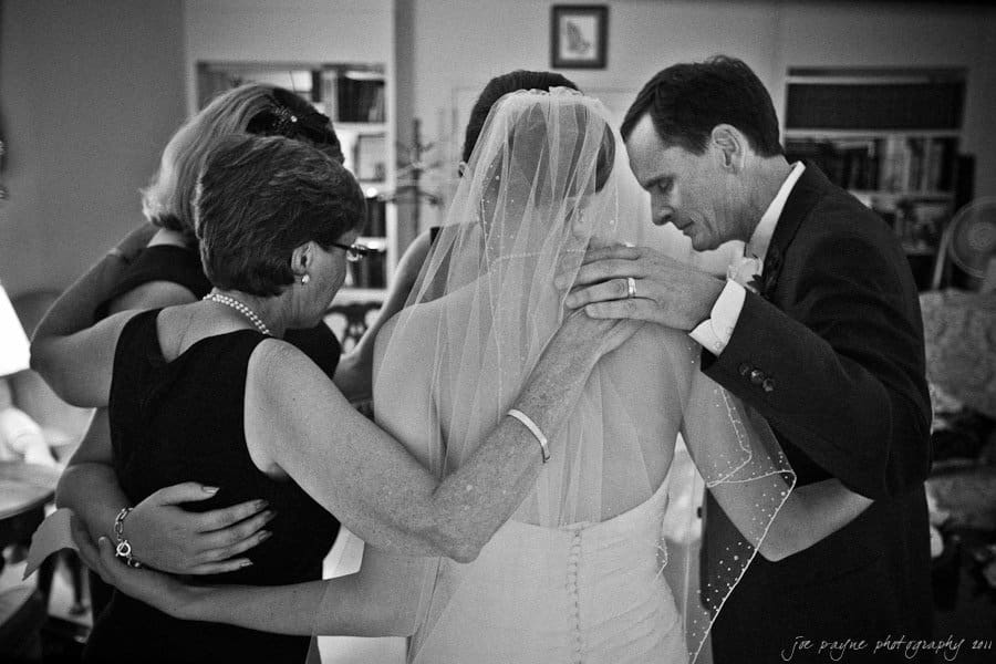 carolina inn weddings | allison and matthew A group of people, including a bride in a wedding dress and veil, are huddled together in a close embrace, sharing an intimate moment before a wedding ceremony in a room filled with bookshelves and cozy furniture.