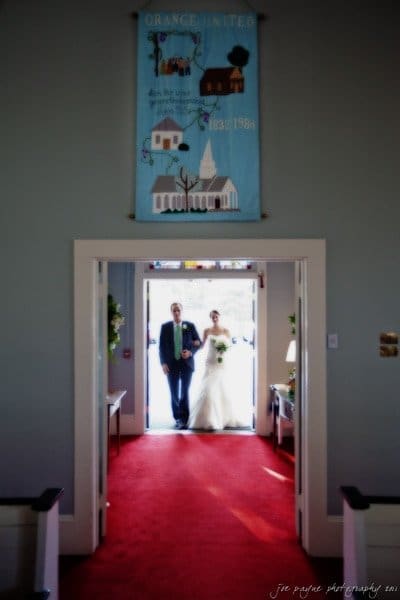 carolina inn weddings | allison and matthew A bride and groom stand in the entrance of a sunlit church with a red carpeted aisle. A banner with text and drawings hangs above the door. Flowers decorate the doorway.