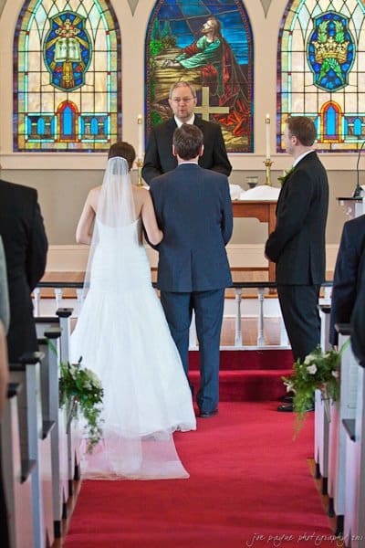 carolina inn weddings | allison and matthew A bride in a white gown with a veil stands at the altar with a groom in a suit, facing a minister in a church. Stained glass windows are visible in the background. Two men in suits stand beside them.