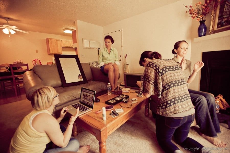 carolina inn weddings | allison and matthew Five women in a living room are gathered for a casual get-together. Some are seated on sofas and the floor, while others interact over a coffee table with a laptop and snacks. The room has neutral tones, a ceiling fan, and a small kitchen visible.