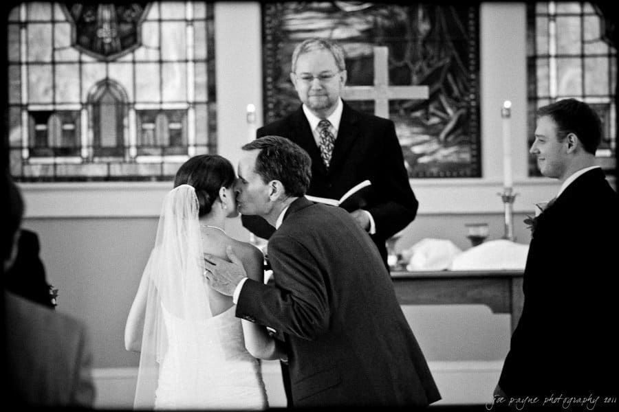 carolina inn weddings | allison and matthew A bride in a white gown and veil is kissed on the cheek by a man in a suit, possibly during a wedding ceremony. They stand in front of a smiling officiant near a stained glass window. Another man in a suit observes the scene.
