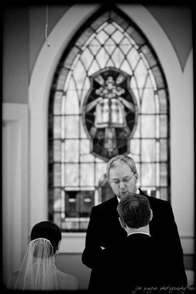 carolina inn weddings | allison and matthew A black and white image of a wedding ceremony inside a church. The officiant stands in front of a stained glass window, with the bride, wearing a veil, and groom facing him. The background features an ornate arched window design.