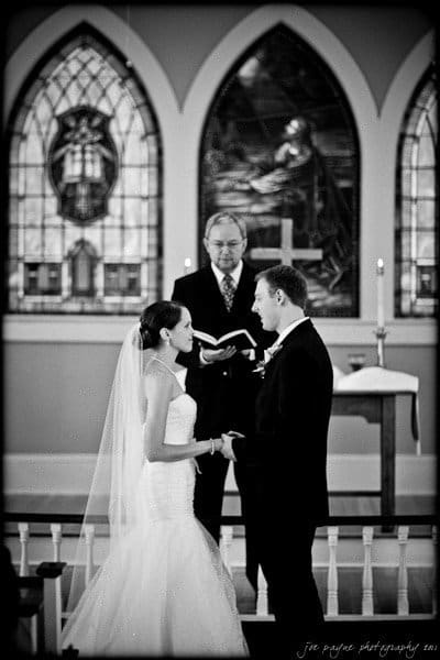 carolina inn weddings | allison and matthew A bride and groom stand facing each other in a church, holding hands during their wedding ceremony. A minister stands behind them, holding a book. Stained glass windows are visible in the background.