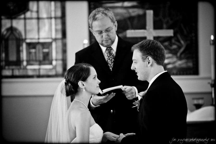 carolina inn weddings | allison and matthew A bride and groom stand facing each other during a wedding ceremony. The bride wears a veil and white dress. The groom is in a suit. A person officiates the ceremony, holding an open book. There is a cross in the background.