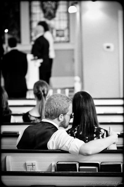 carolina inn weddings | allison and matthew Black and white photo of a wedding ceremony in a church. A couple sits in the pews, with the man wrapping his arm around the woman. The bride and groom are at the altar in the background.