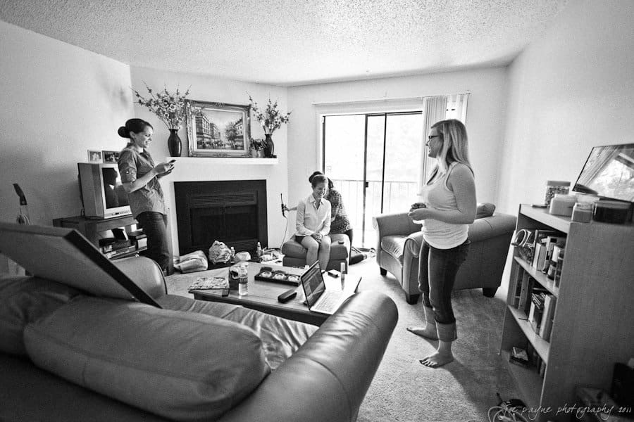 A black and white photo of three people in a living room. One is standing near a fireplace holding a phone, another sits on a couch with a laptop, and the third stands barefoot holding a mug. The room is cozy with a window letting in natural light.